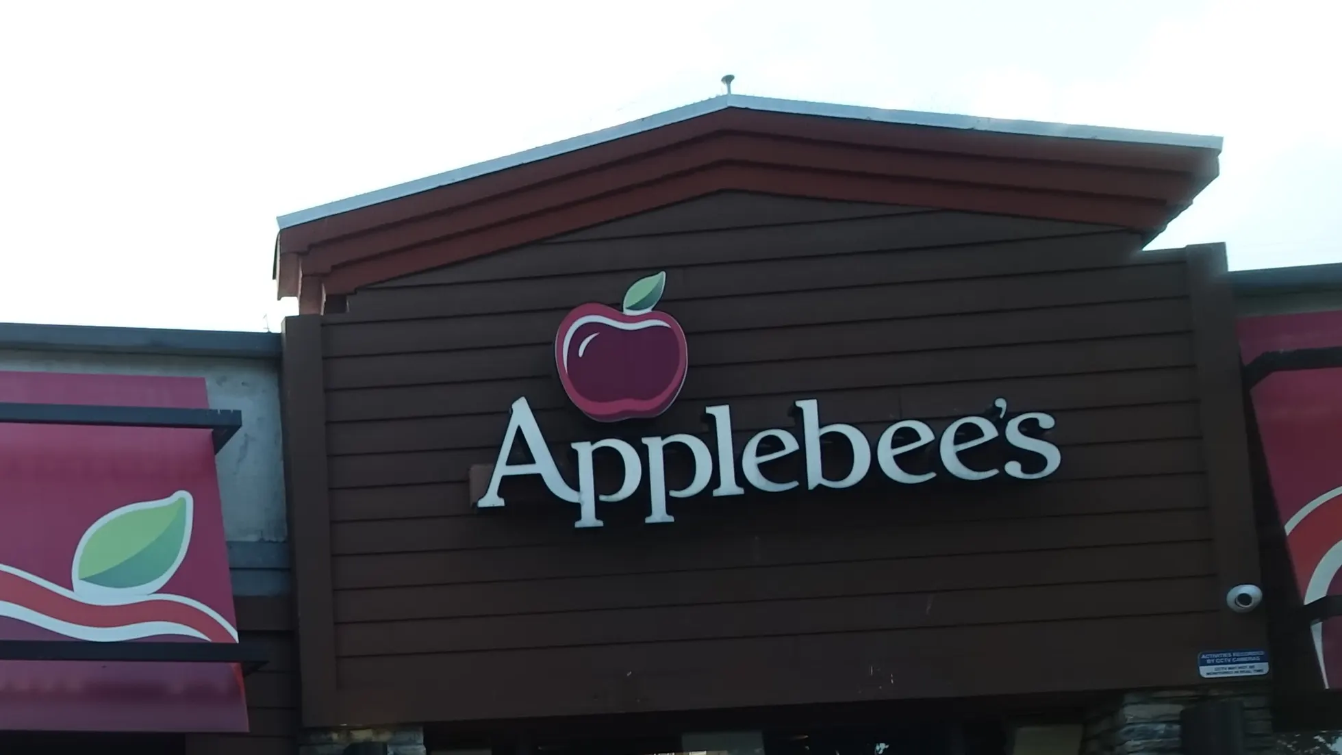 Parte superior de fachada marrom com letreiro Applebee's e símbolo de maçã vermelha, telhado inclinado e parte de toldo vermelho à esquerda
