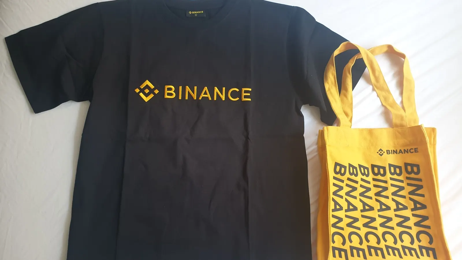 Camiseta preta e bolsa amarela com a marca Binance sobre uma superfície branca