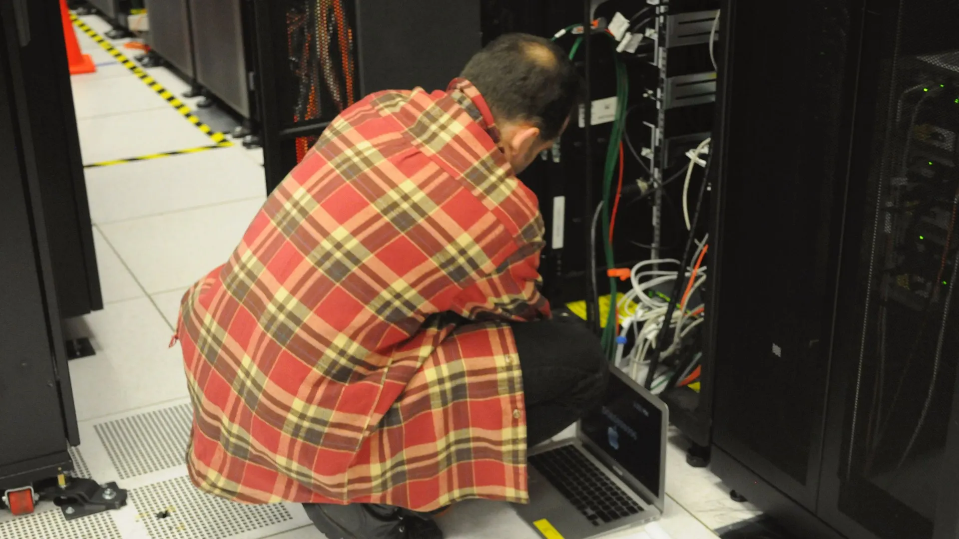 Pessoa de costas com camisa xadrez vermelha inclinada diante de rack aberto, com cabos e notebook sobre o piso