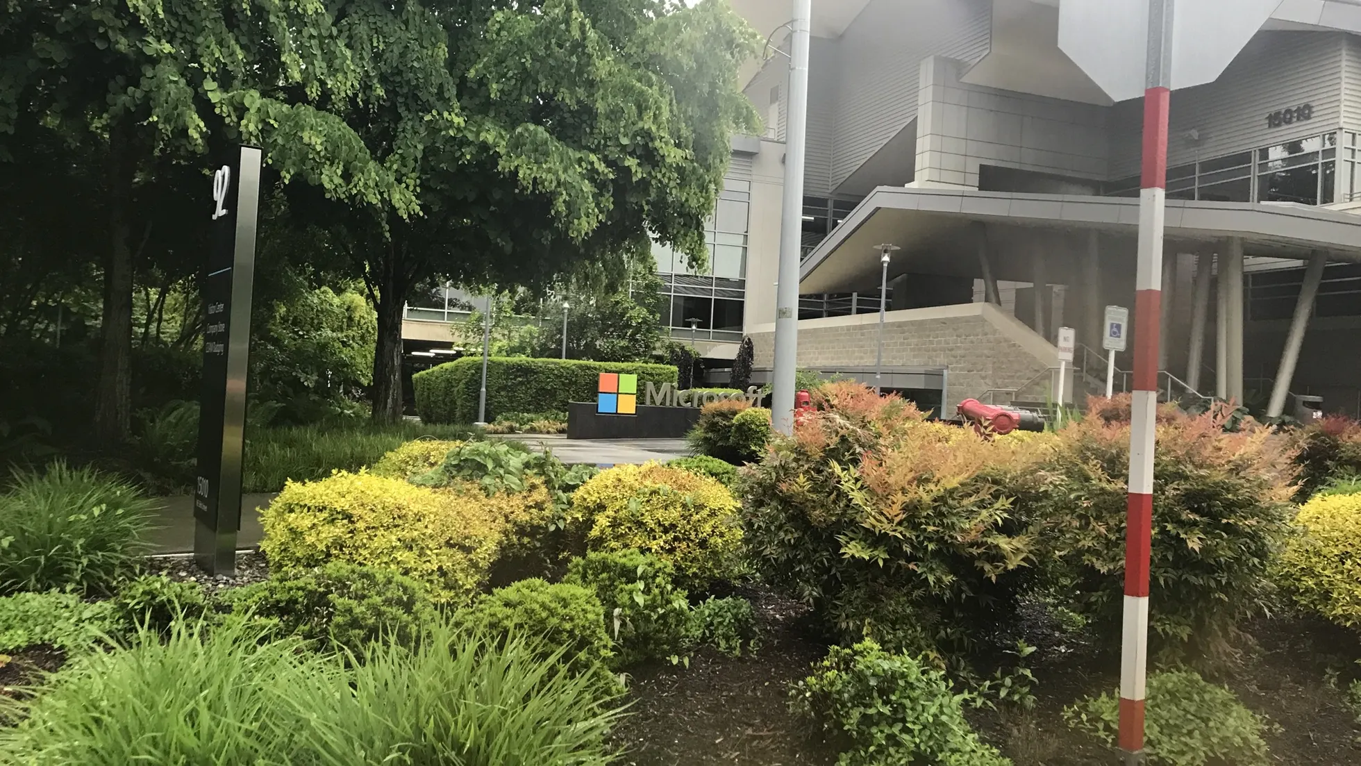 Entrada de prédio com letreiro Microsoft entre arbustos, árvores à esquerda, placa preta com número 92 e poste branco e vermelho à direita