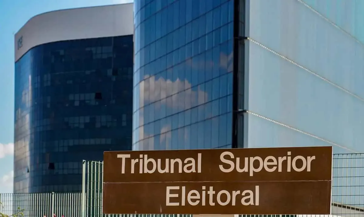 Placa de identificação em madeira com a inscrição Tribunal Superior Eleitoral em frente a um prédio com fachada de vidro azul