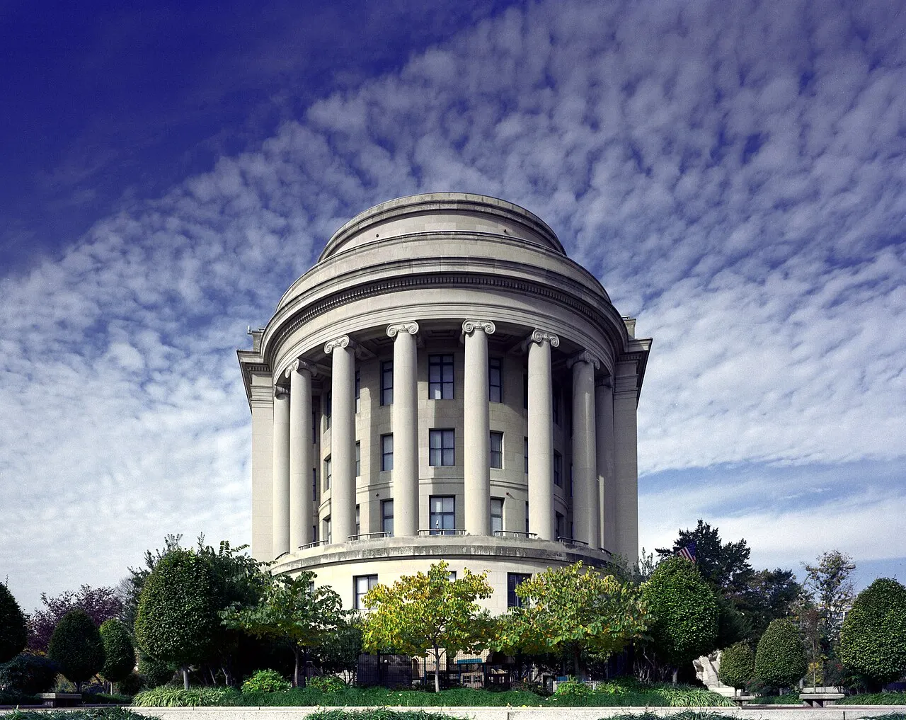 Fachada semicircular do Edifício da Federal Trade Commission em Washington, com colunas jônicas clássicas de pedra clara no topo, janelas escuras dispostas em vários andares, copa de árvores com folhagem verde e amarelada na base e céu azul coberto por nuvens brancas onduladas