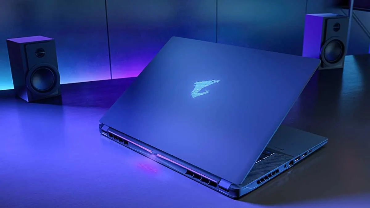 Notebook gamer Gigabyte Aorus Master 16 repousado sobre uma mesa com luz azulada, exibindo a tampa com o logotipo brilhante da marca e caixas de som ao fundo