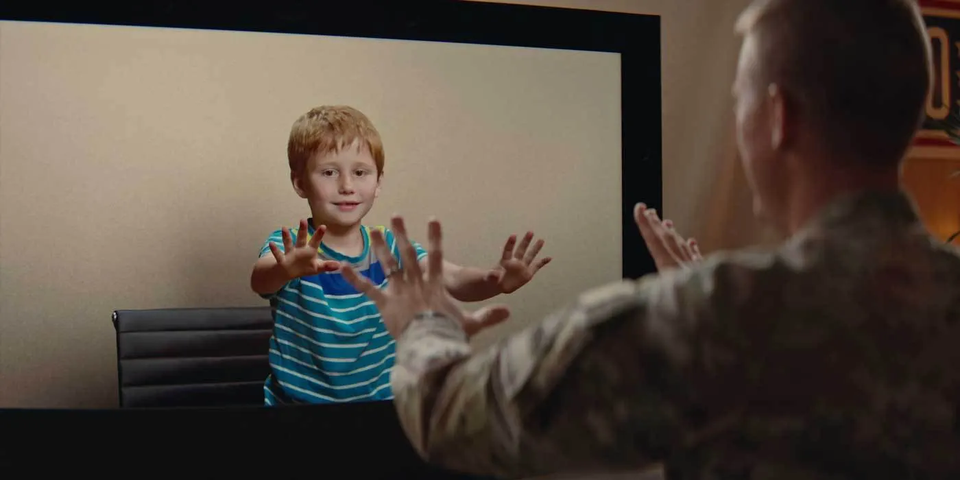 Criança de camiseta listrada estende as mãos para a tela de uma cabine de videochamada 3D, enquanto um militar de costas estende as mãos na direção dela