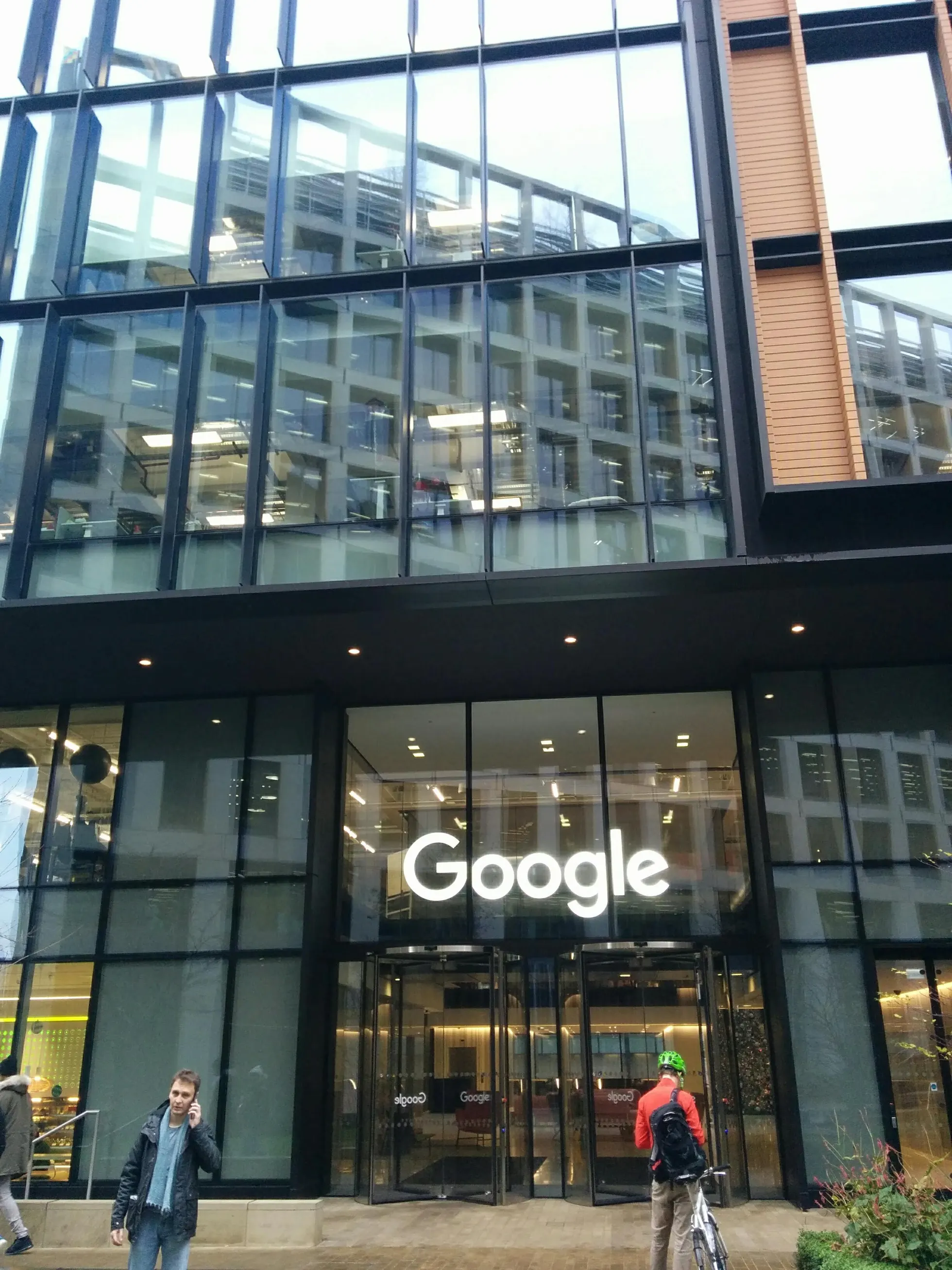 Entrada envidraçada de edifício comercial moderno com letreiro luminoso branco Google sobre portas giratórias de vidro. À esquerda, um pedestre de casaco escuro, cachecol azul claro e calça jeans fala ao celular. À direita, um ciclista de costas com jaqueta vermelha, capacete verde-limão e mochila preta segura uma bicicleta prateada. Acima da entrada, a fachada envidraçada reflete um prédio com janelas quadradas e traz acabamento lateral em painéis verticais de madeira clara