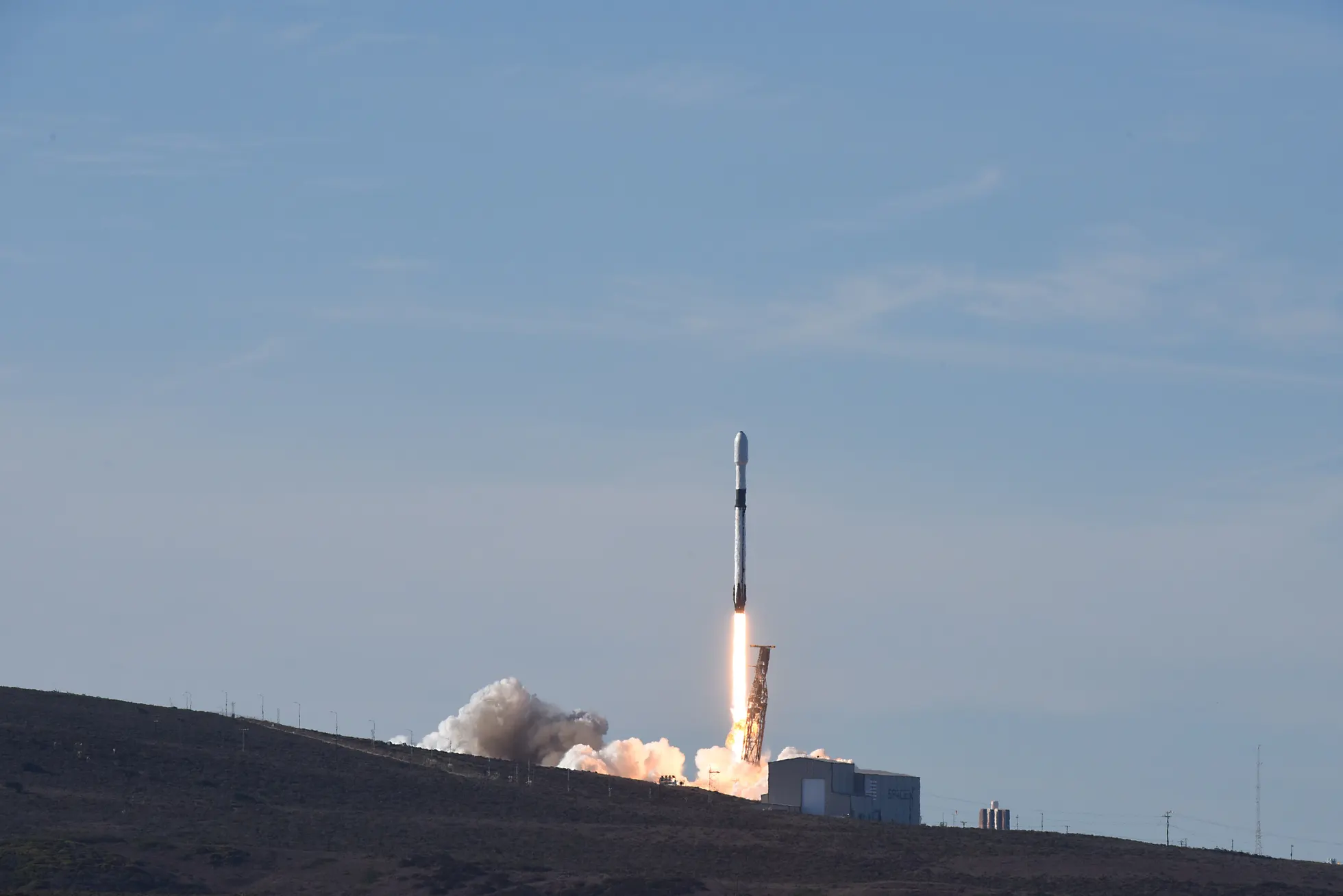 Foguete Falcon 9 branco e preto em ascensão vertical após o lançamento, expelindo labaredas alaranjadas e fumaça branca densa sobre a base costeira, com colina escura em primeiro plano e céu azul ao fundo