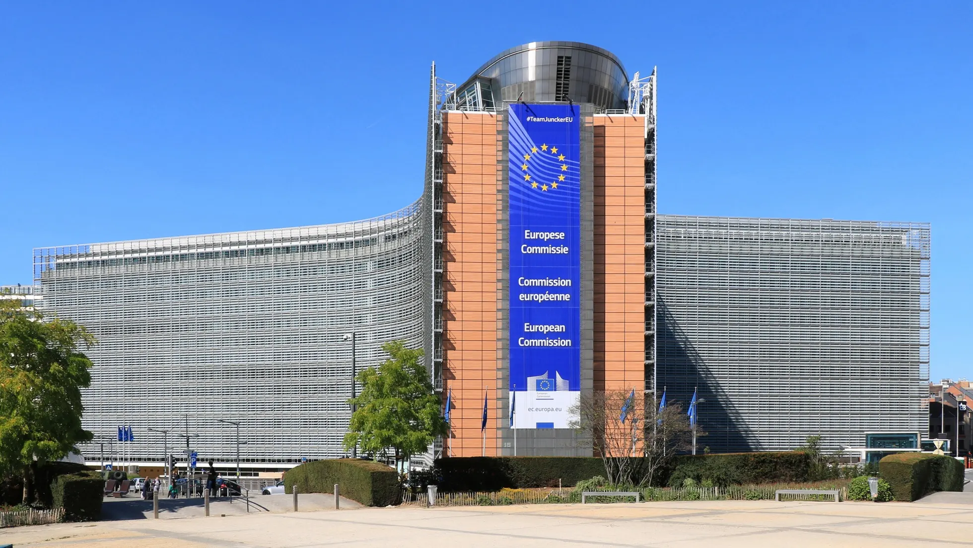 Prédio com fachada de vidro e estrutura central alaranjada, faixa azul com estrelas amarelas e European Commission, diante de praça com árvores sob céu azul