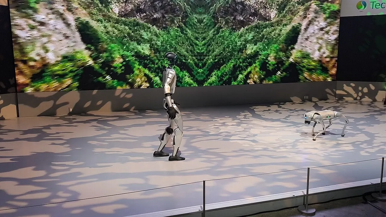 Robô humanoide prateado de perfil e robô quadrúpede branco em palco, diante de telão com imagem verde de vegetação
