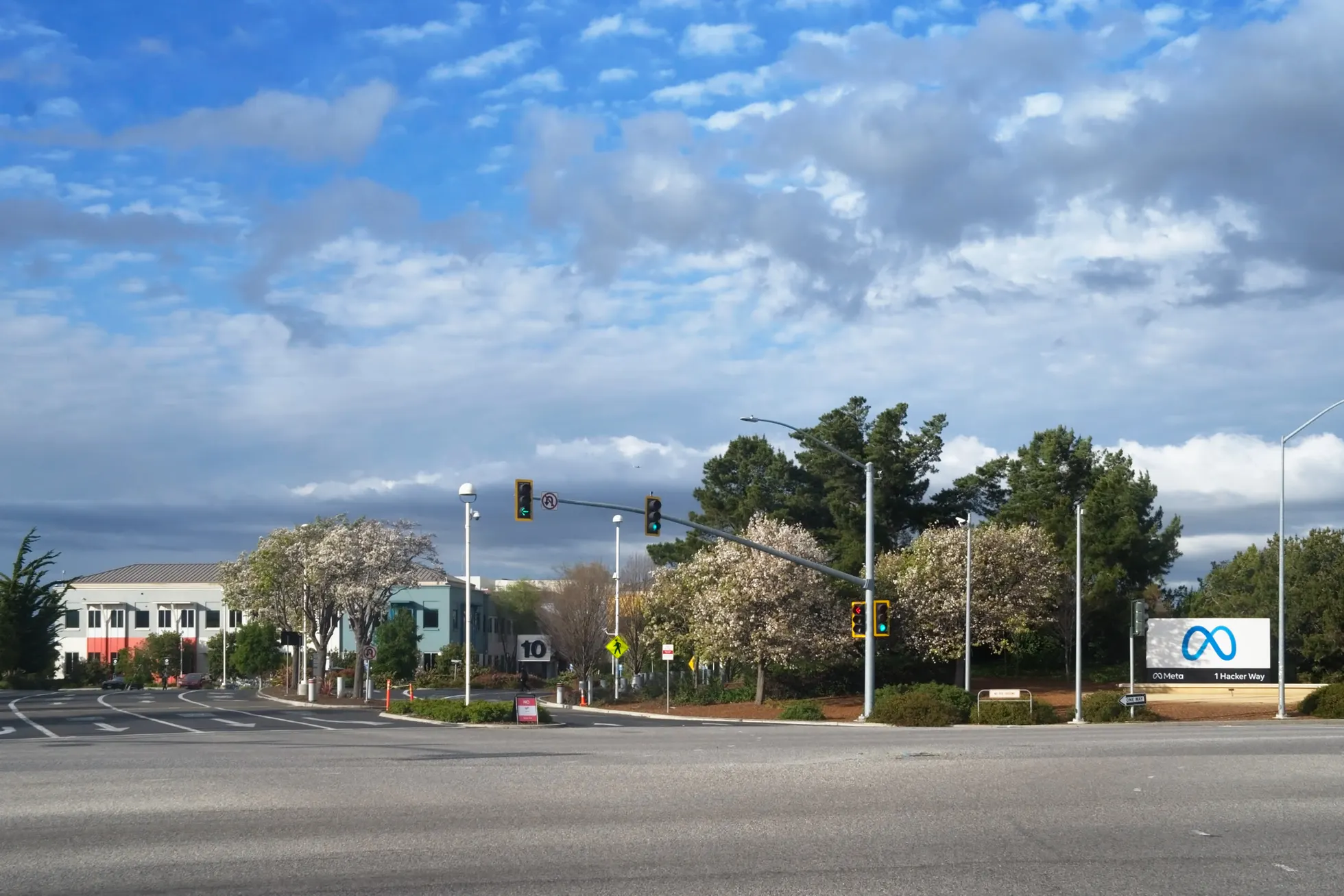 Letreiro branco tridimensional com as letras META sobre a grama, em frente a um prédio de vidro que mostra o símbolo do infinito e a palavra META, com uma avenida de várias faixas, carros, semáforos, árvores e céu azul com nuvens
