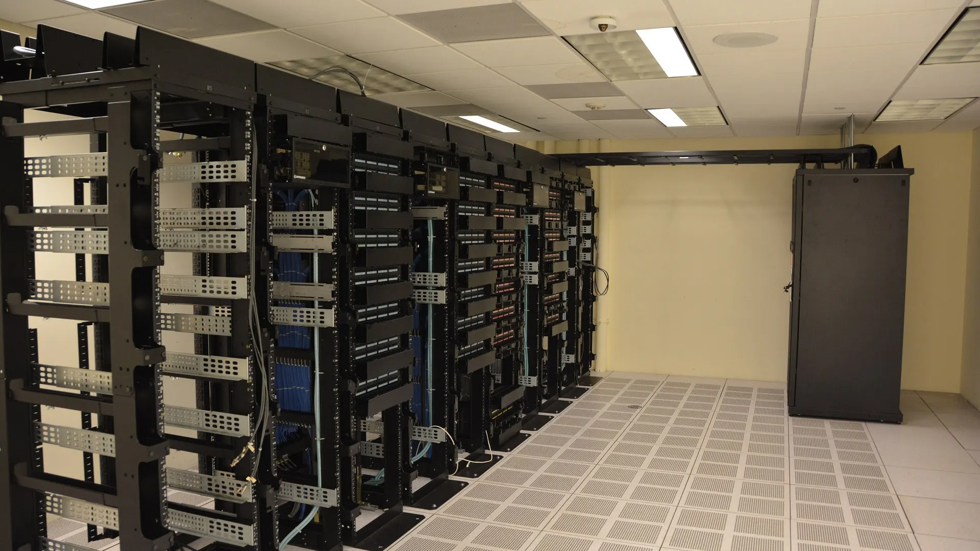 Sala de data center com fileiras de racks de servidores pretos, cabos azuis e piso elevado perfurado