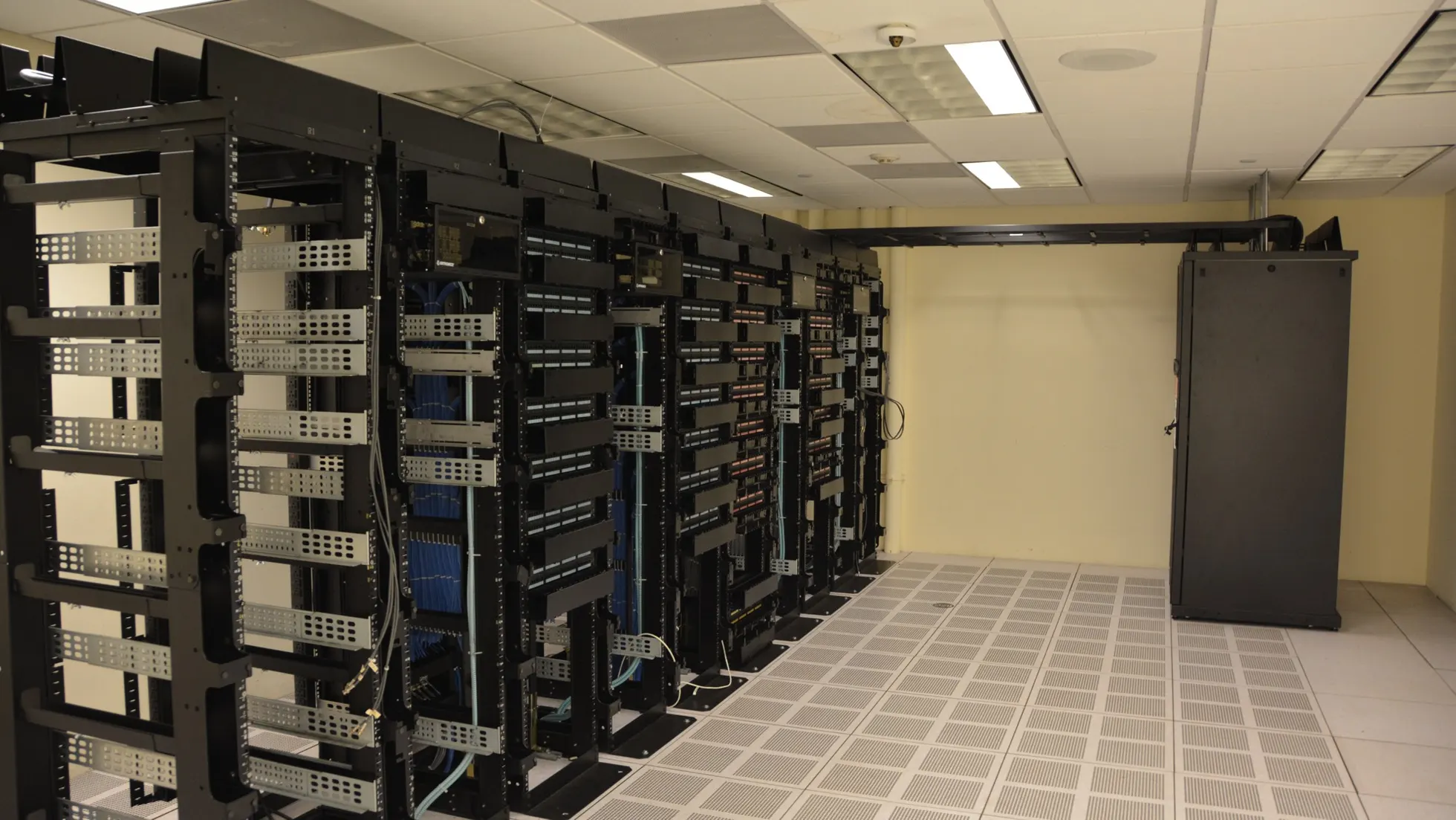 Fileiras de racks pretos com equipamentos e cabos em sala clara com piso ventilado e luminárias no teto