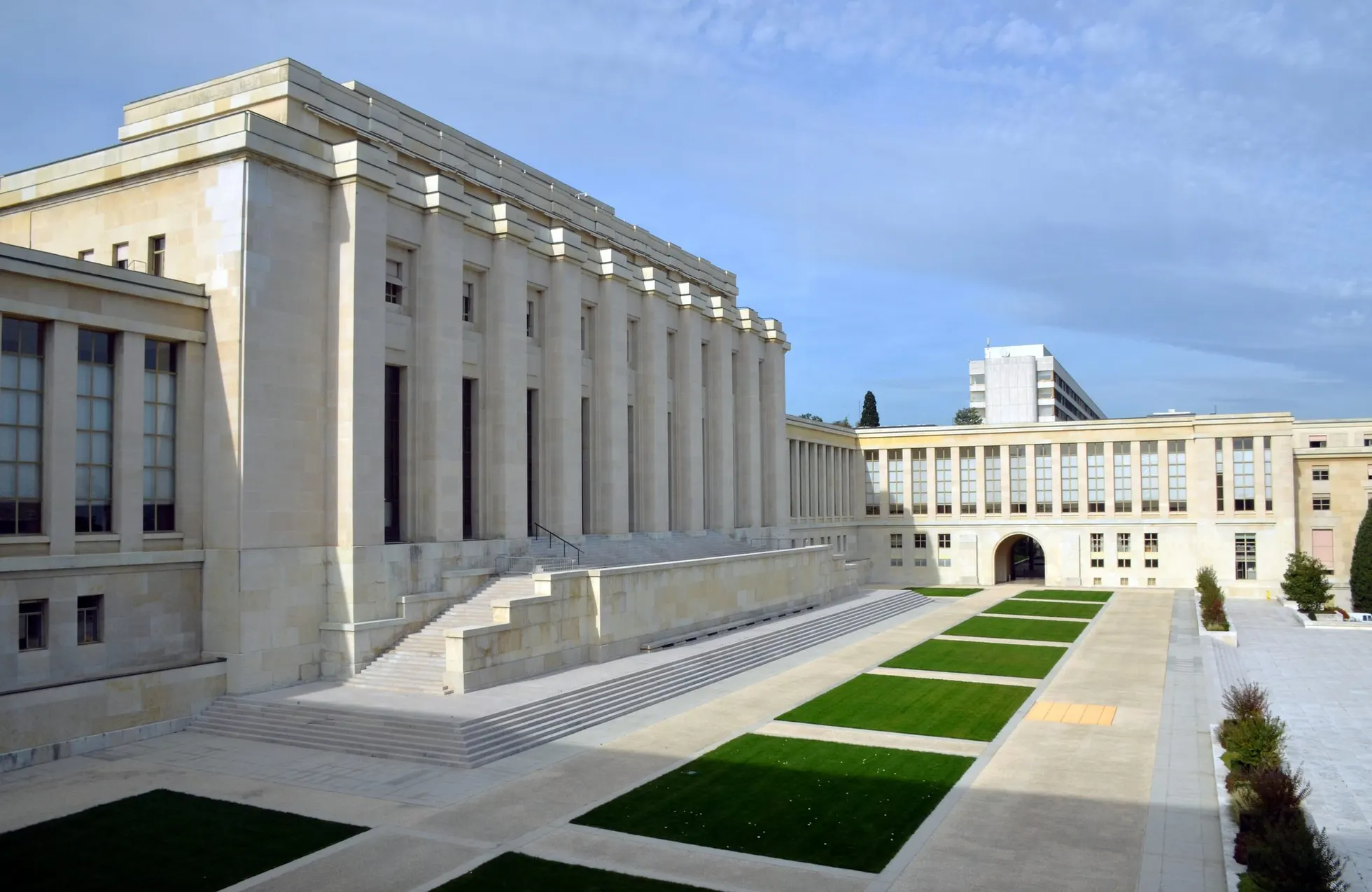 Fachada do Palais des Nations, sede da ONU em Genebra: prédio de pedra clara com colunas altas, escadaria lateral e pátio com canteiros retangulares de grama, sob céu azul