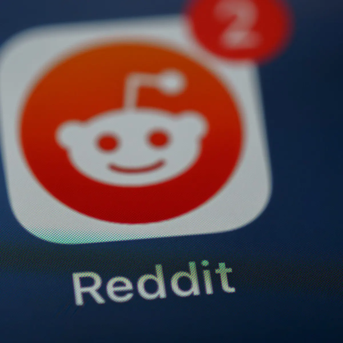 Ícone do aplicativo Reddit exibido em tela de smartphone com pixels visíveis, destacando a cabeça branca do mascote Snoo sobre círculo vermelho-alaranjado dentro de quadrado branco de cantos arredondados, distintivo de notificação vermelho com o número 2 no canto superior direito e a palavra Reddit em letras brancas logo abaixo