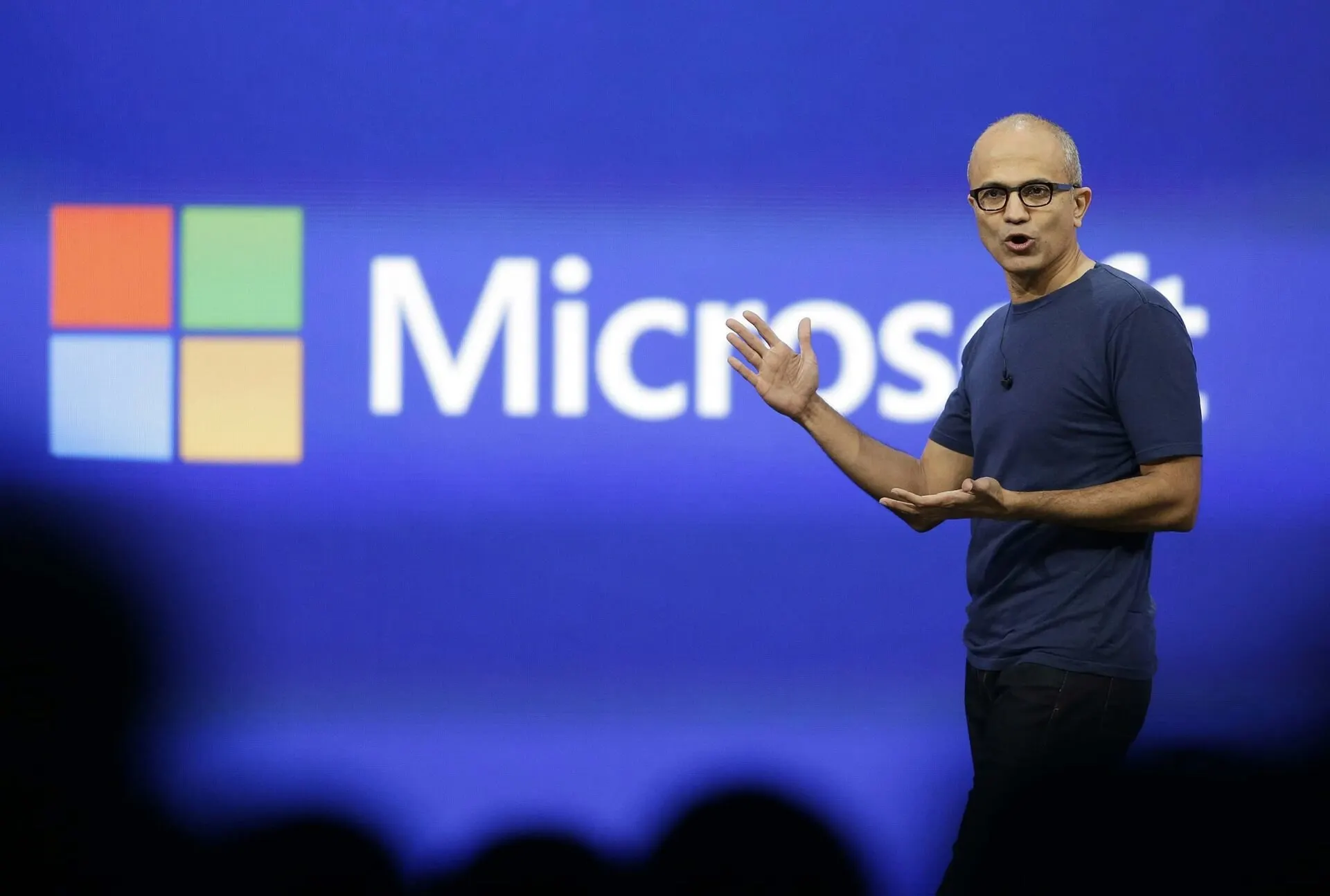 Satya Nadella vestindo camiseta azul escura e óculos, com os braços abertos gesticulando no palco diante de um telão azul com o logotipo da Microsoft
