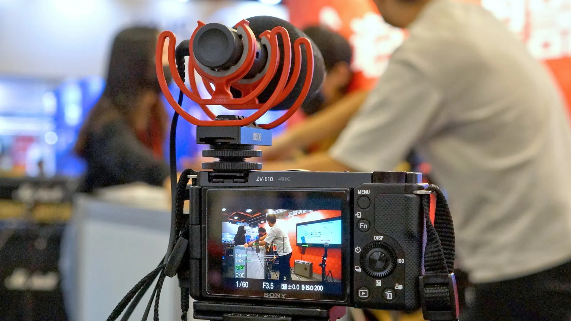 Câmera Sony ZV-E10 em tripé com microfone de vídeo vermelho e preto na sapata, usada como imagem ilustrativa; não é o ECM-AX10