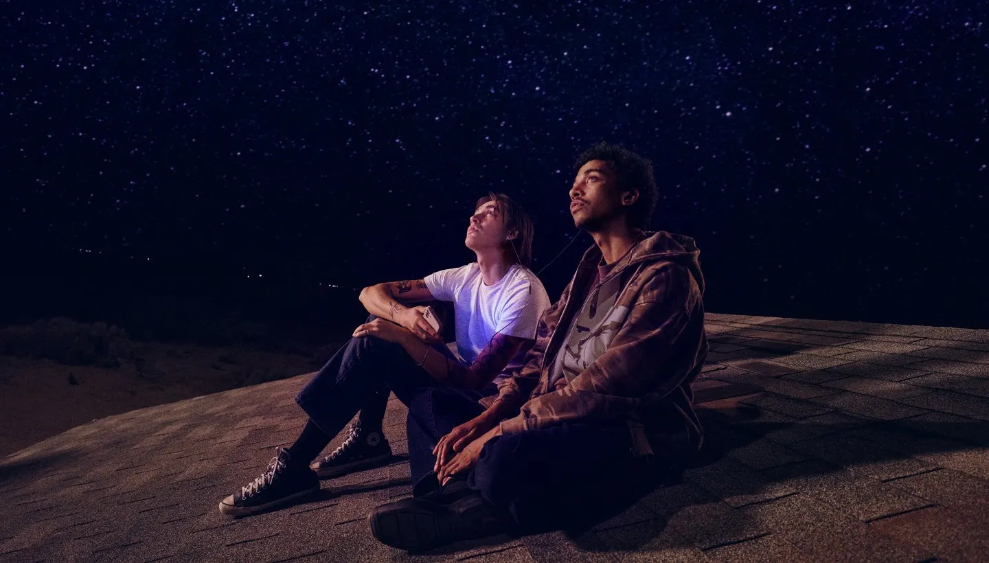Dois jovens sentados no telhado de uma casa durante a noite sob céu estrelado, com um deles segurando um celular conectado a fones de ouvido.