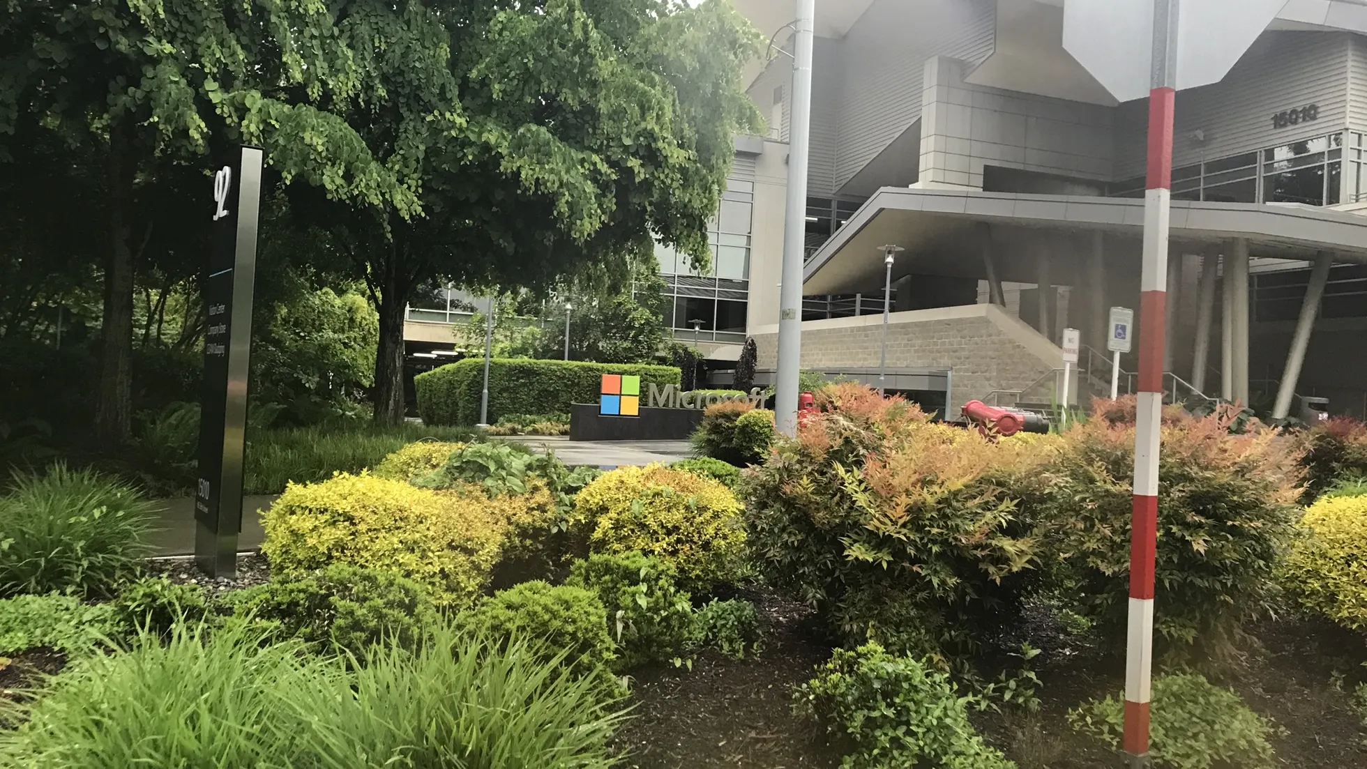Jardim com arbustos e árvores diante de prédio cinza, placa com logotipo Microsoft ao centro e postes claros na frente