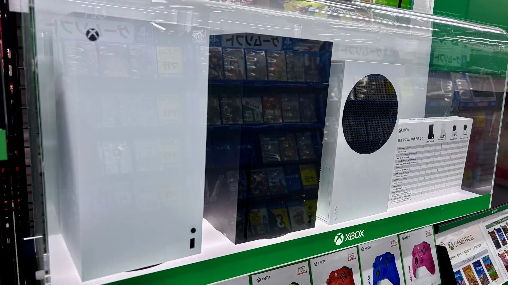 Vitrine de loja com um Xbox Series X branco e um Xbox Series S ao lado, acima de caixas de controles e do selo Xbox em verde
