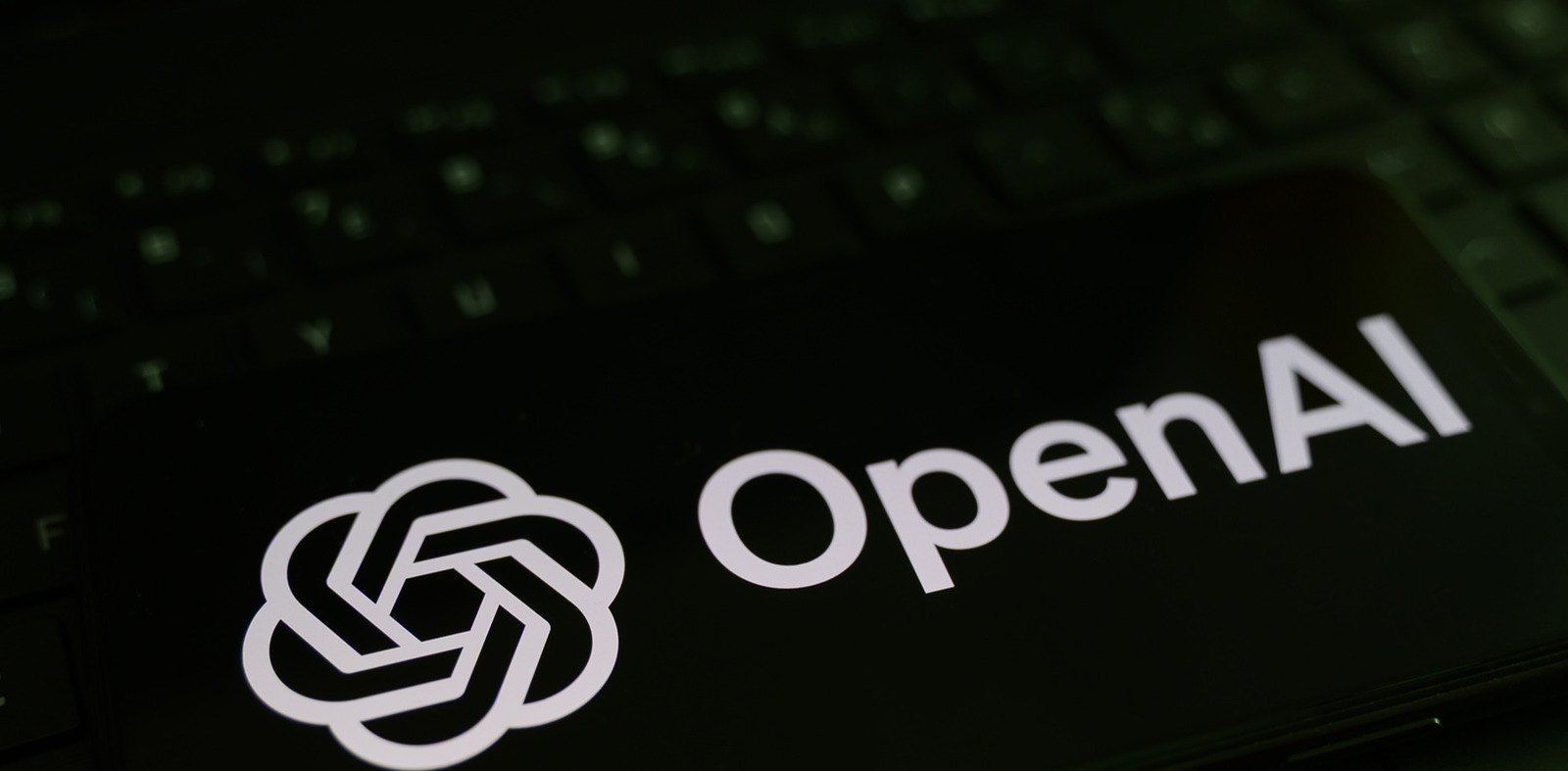 Smartphone com o logotipo branco da OpenAI em destaque na tela preta, apoiado sobre o teclado escuro de um computador portátil.