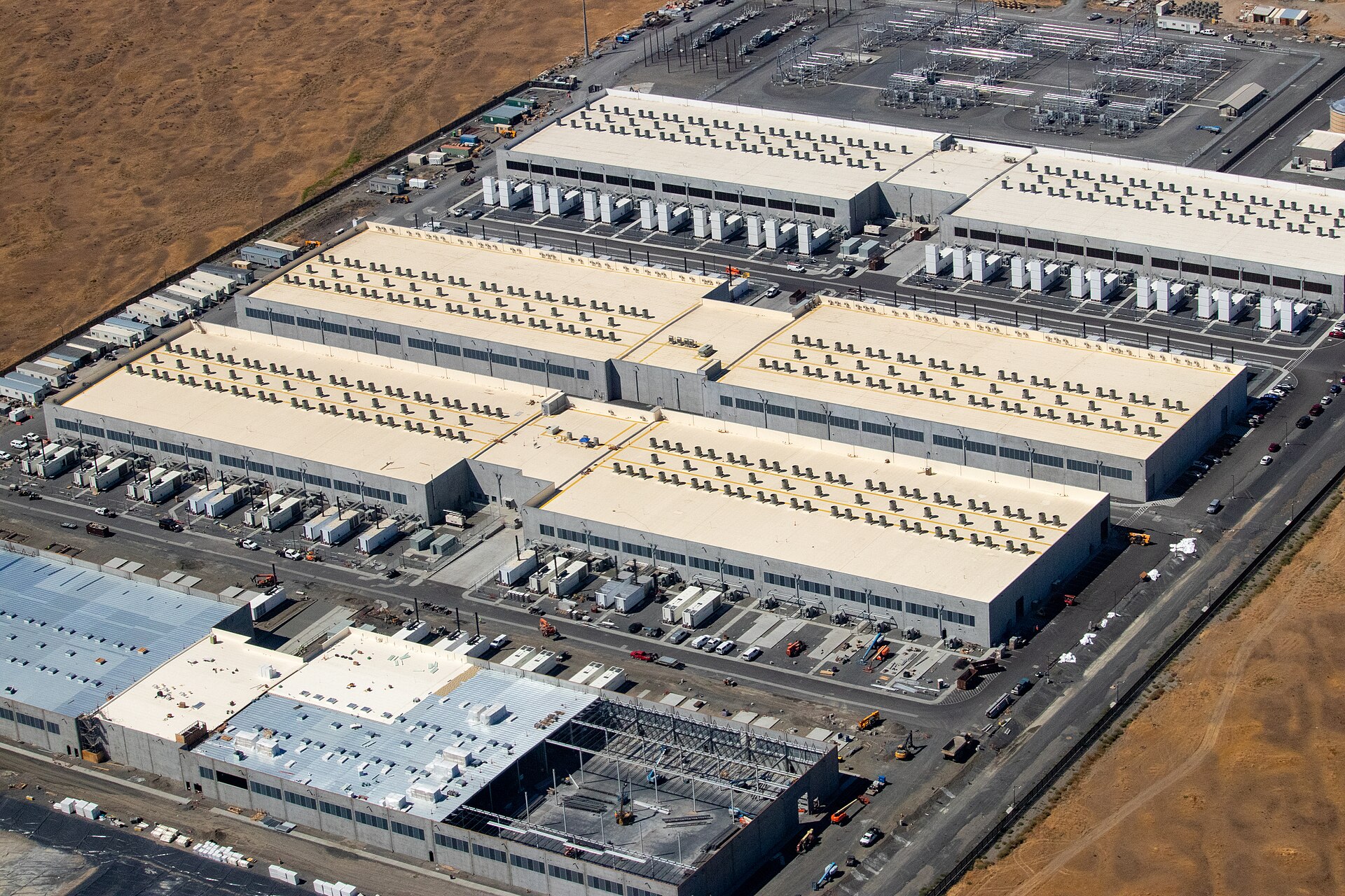 Vista aérea de complexo de data centers da Amazon Web Services em terreno árido de terra marrom clara, com três galpões industriais cinzas compridos com fileiras regulares de exaustores de ar no teto branco, um quarto galpão com estrutura metálica em obras no canto inferior esquerdo e subestação elétrica com torres metálicas e transformadores no topo