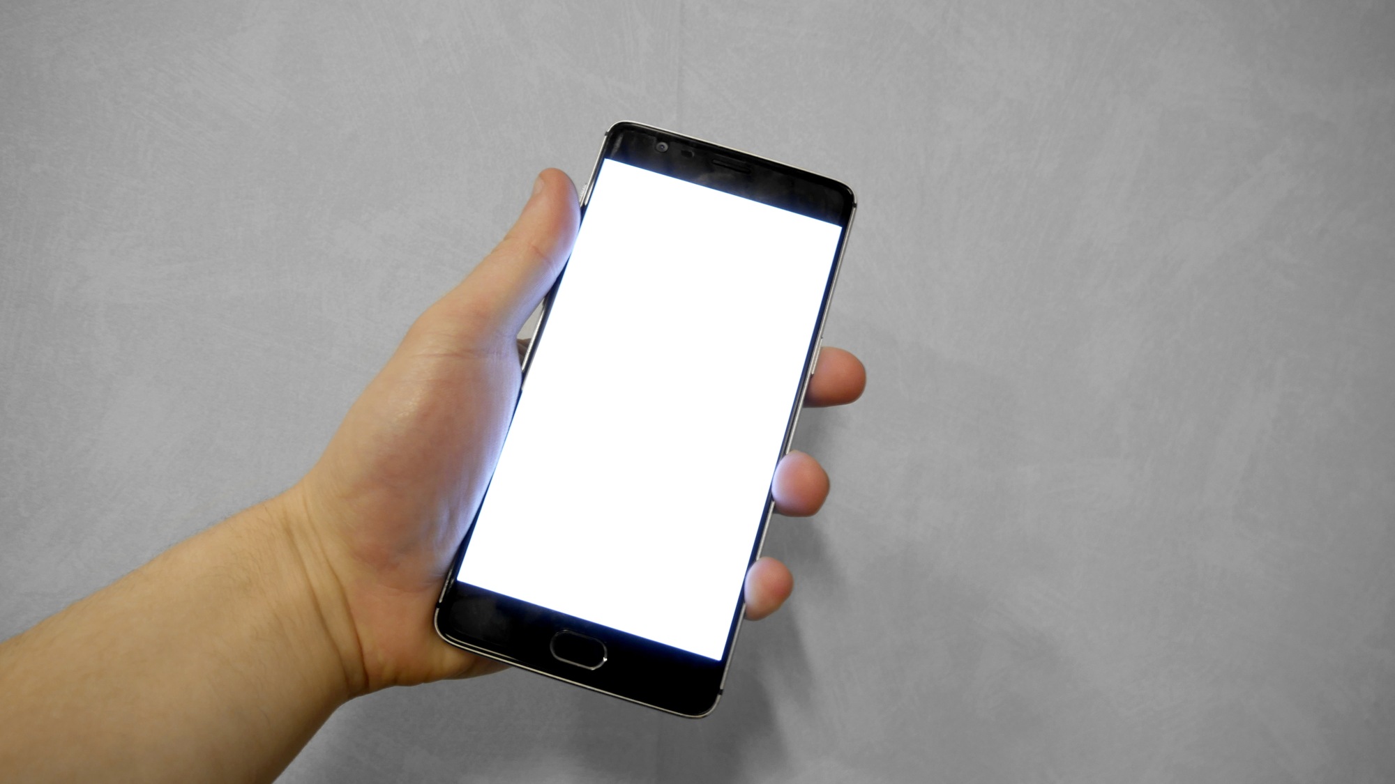 Mão segurando celular preto com tela branca acesa diante de uma parede cinza