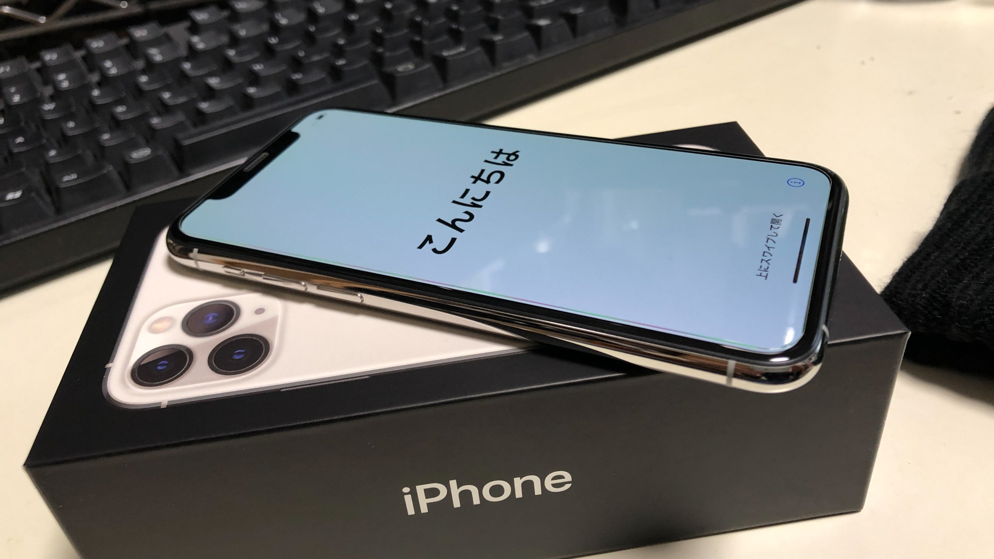 iPhone com tela clara e caracteres japoneses sobre caixa preta, com teclado preto e mesa clara ao fundo