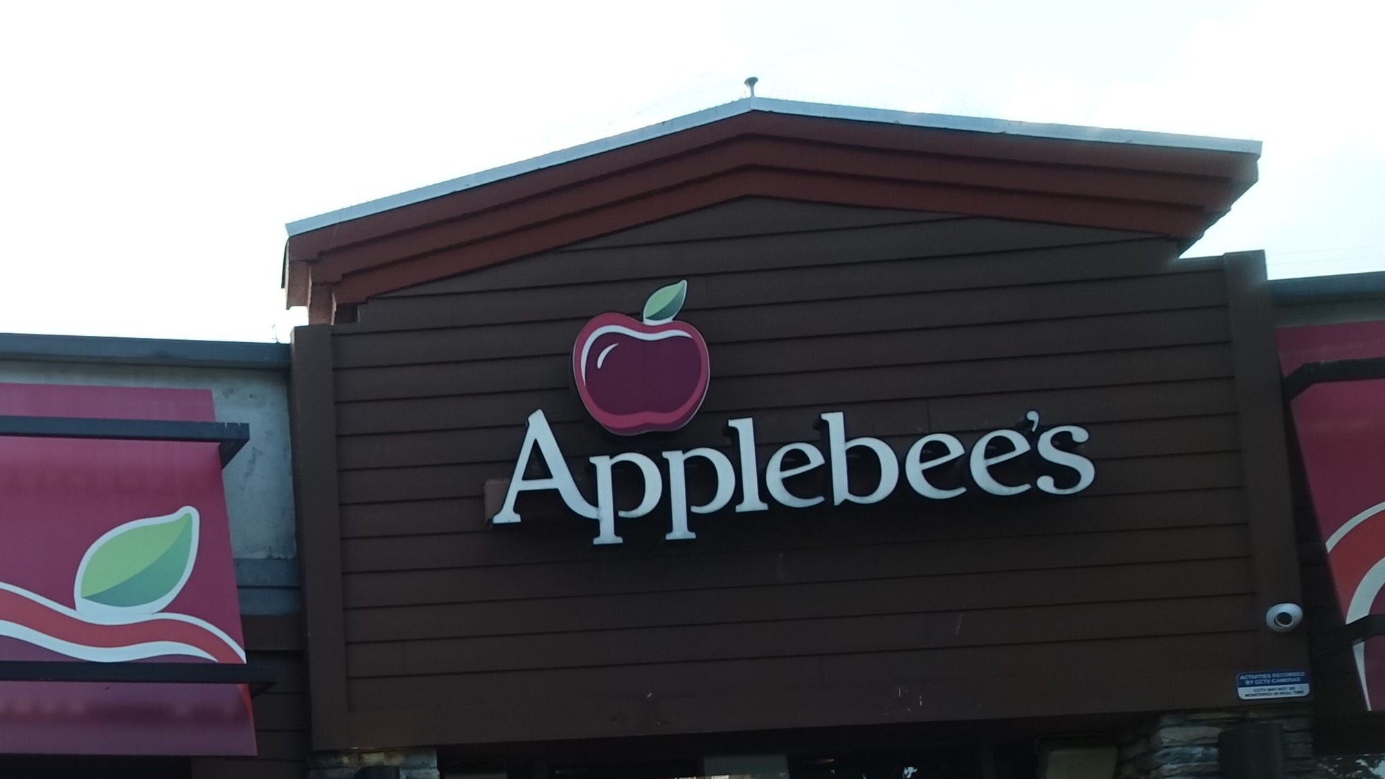 Parte superior de fachada marrom com letreiro Applebee's e símbolo de maçã vermelha, telhado inclinado e parte de toldo vermelho à esquerda