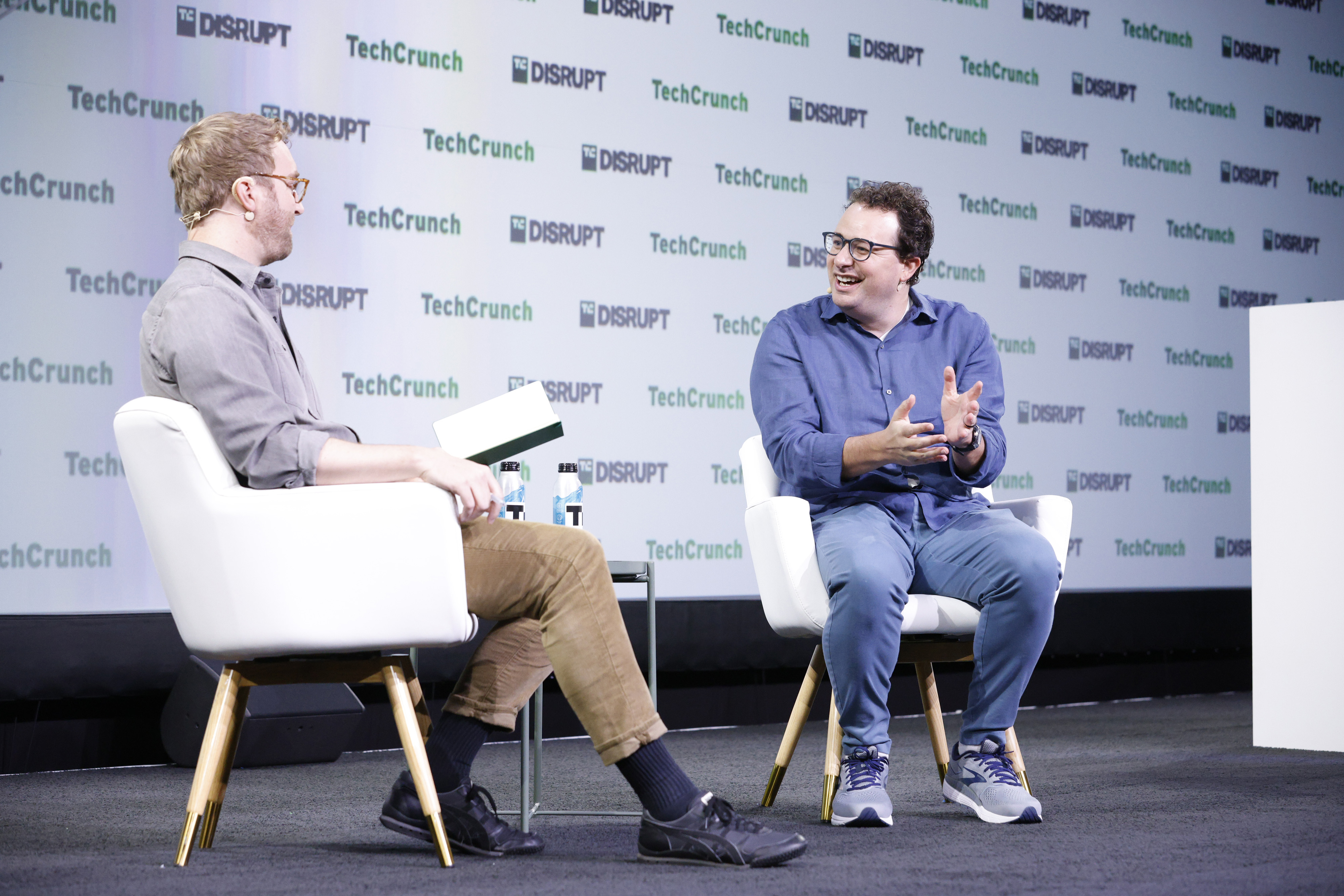 Dario Amodei, de óculos e camisa azul, sentado numa poltrona branca, sorri e gesticula enquanto fala com o entrevistador à esquerda, diante do painel branco com os logotipos TechCrunch e Disrupt
