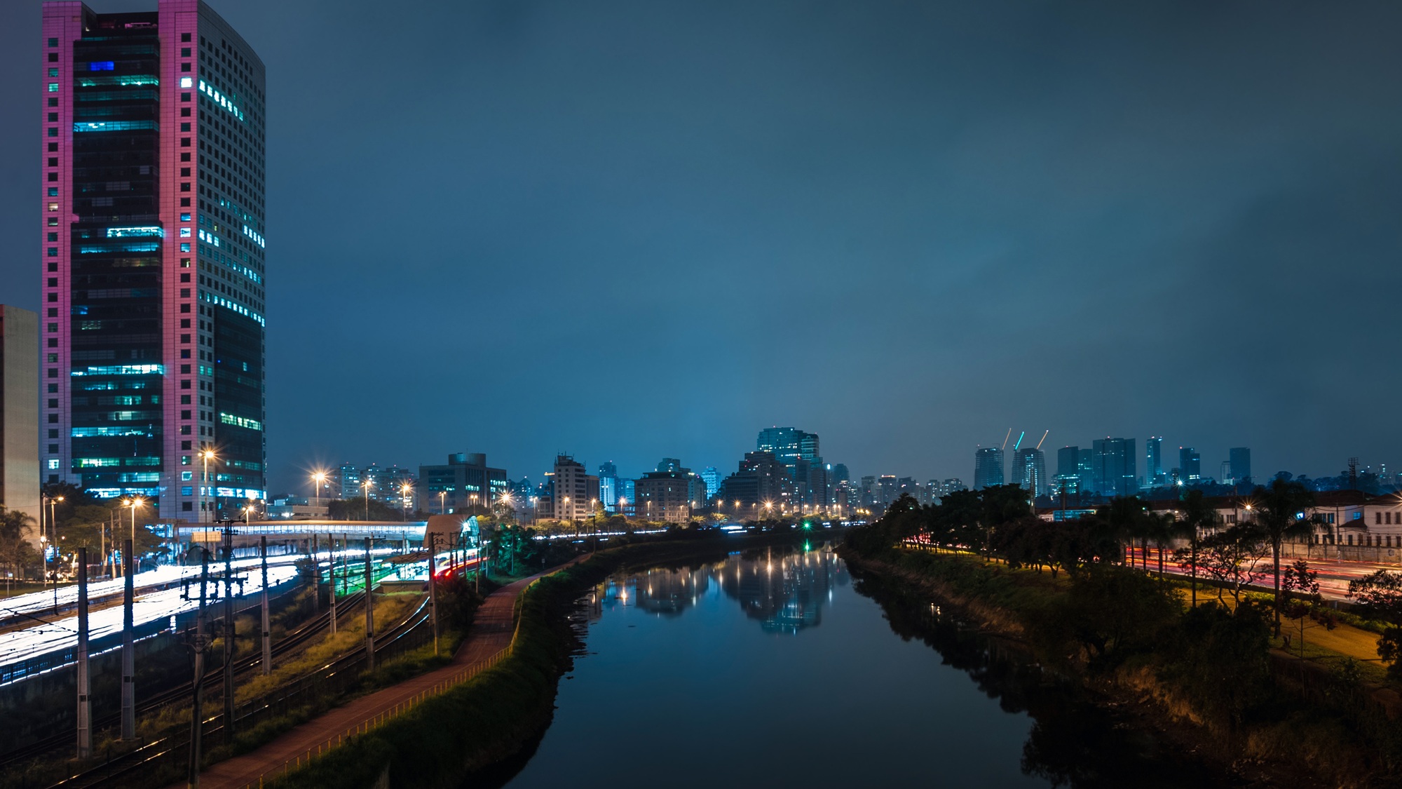 Rio entre margens arborizadas à noite, com prédios iluminados ao fundo, torre alta à esquerda e rastros de luz junto aos trilhos
