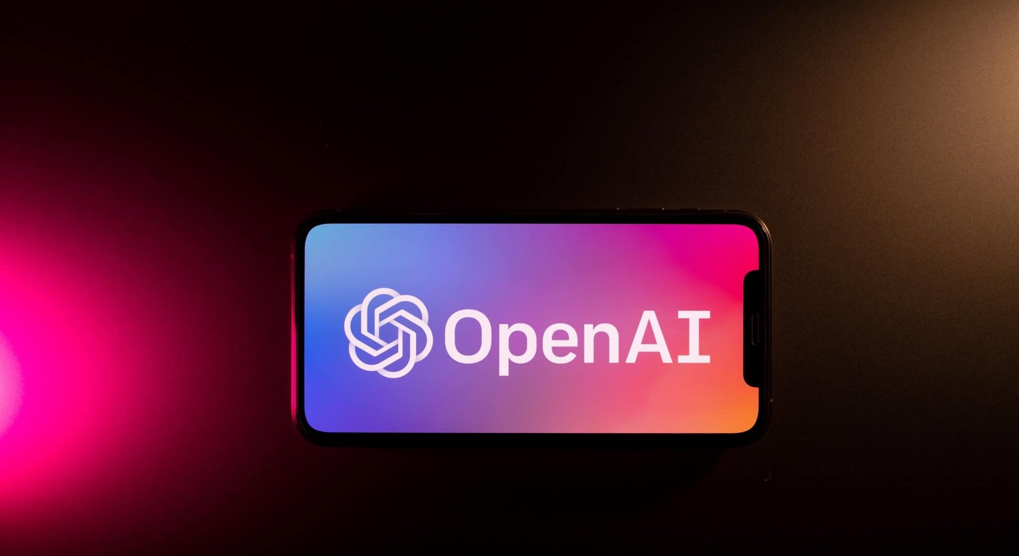 Smartphone posicionado na horizontal exibindo o logotipo da OpenAI em fundo escuro com feixe de iluminação rosa à esquerda