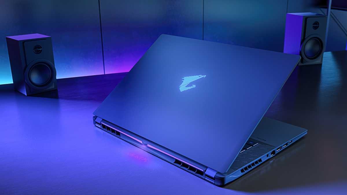 Notebook gamer Gigabyte Aorus Master 16 repousado sobre uma mesa com luz azulada, exibindo a tampa com o logotipo brilhante da marca e caixas de som ao fundo
