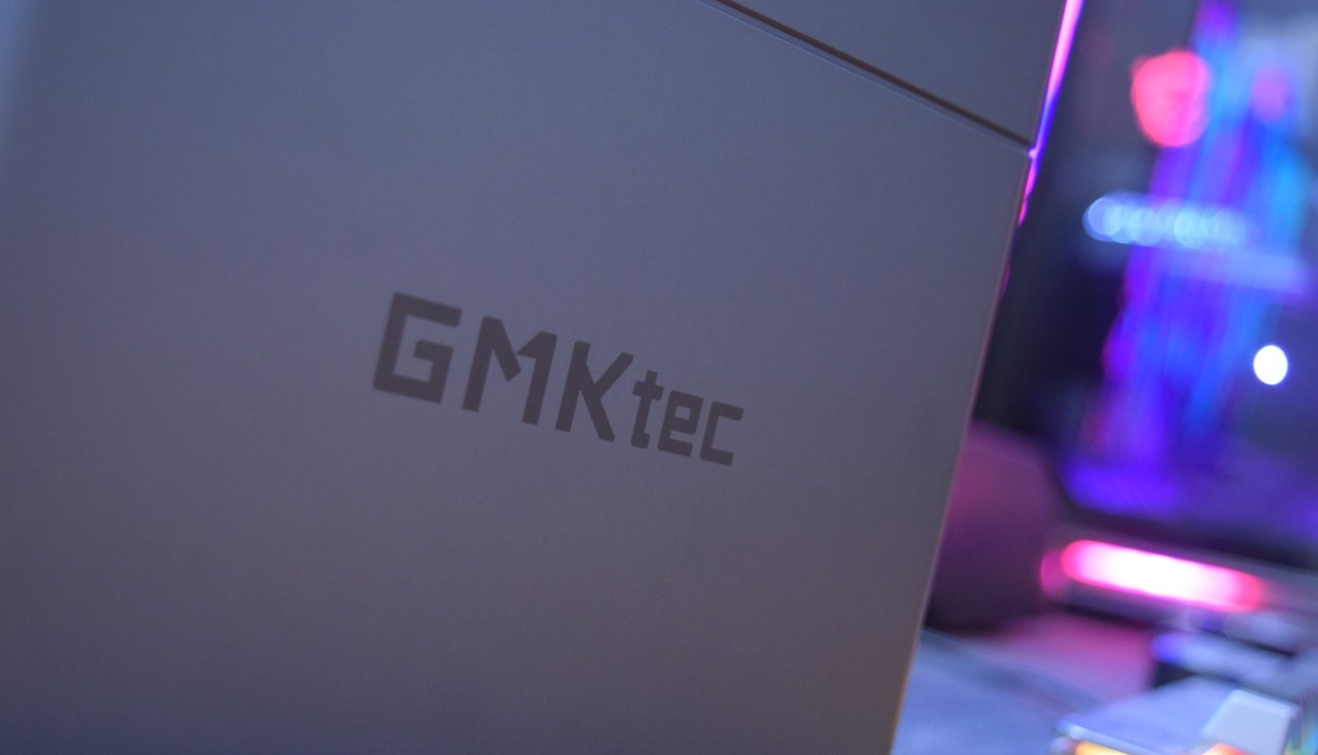 Fotografia em primeiro plano mostrando a logo da marca GMKtec gravada na carcaça metálica de um mini PC, com iluminação em tons de azul e rosa ao fundo.