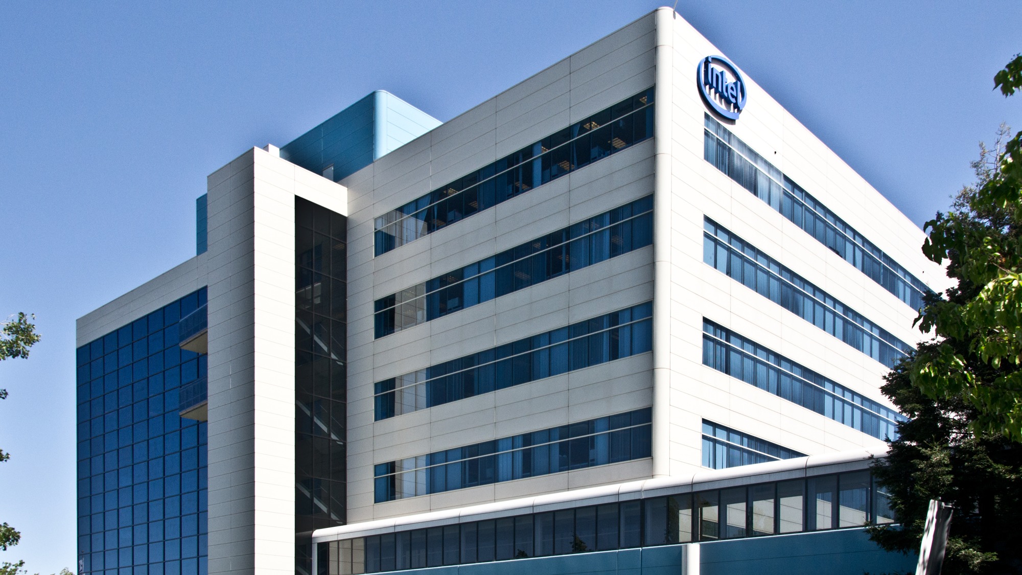 Prédio de fachada branca e janelas azuis com marca Intel na parte superior, árvores nas bordas e céu azul