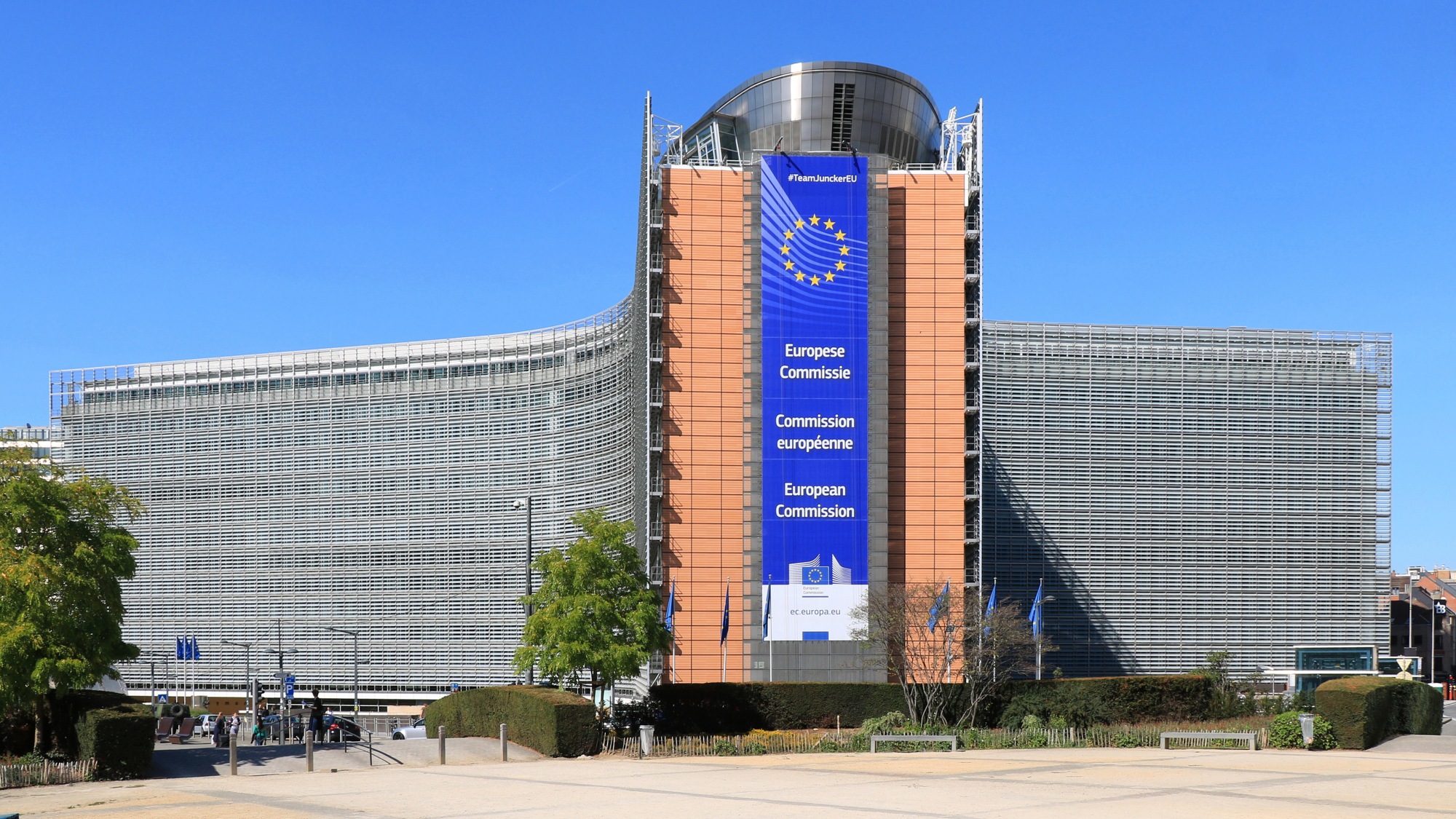 Prédio com fachada de vidro e estrutura central alaranjada, faixa azul com estrelas amarelas e European Commission, diante de praça com árvores sob céu azul