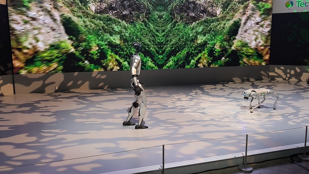 Robô humanoide prateado de perfil e robô quadrúpede branco em palco, diante de telão com imagem verde de vegetação