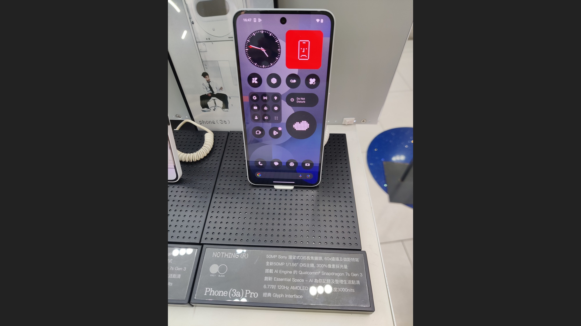 Celular em pé sobre bancada de exposição, com tela roxa de ícones e relógio, cabo branco à esquerda e placa Phone (3a) Pro abaixo, entre faixas escuras laterais