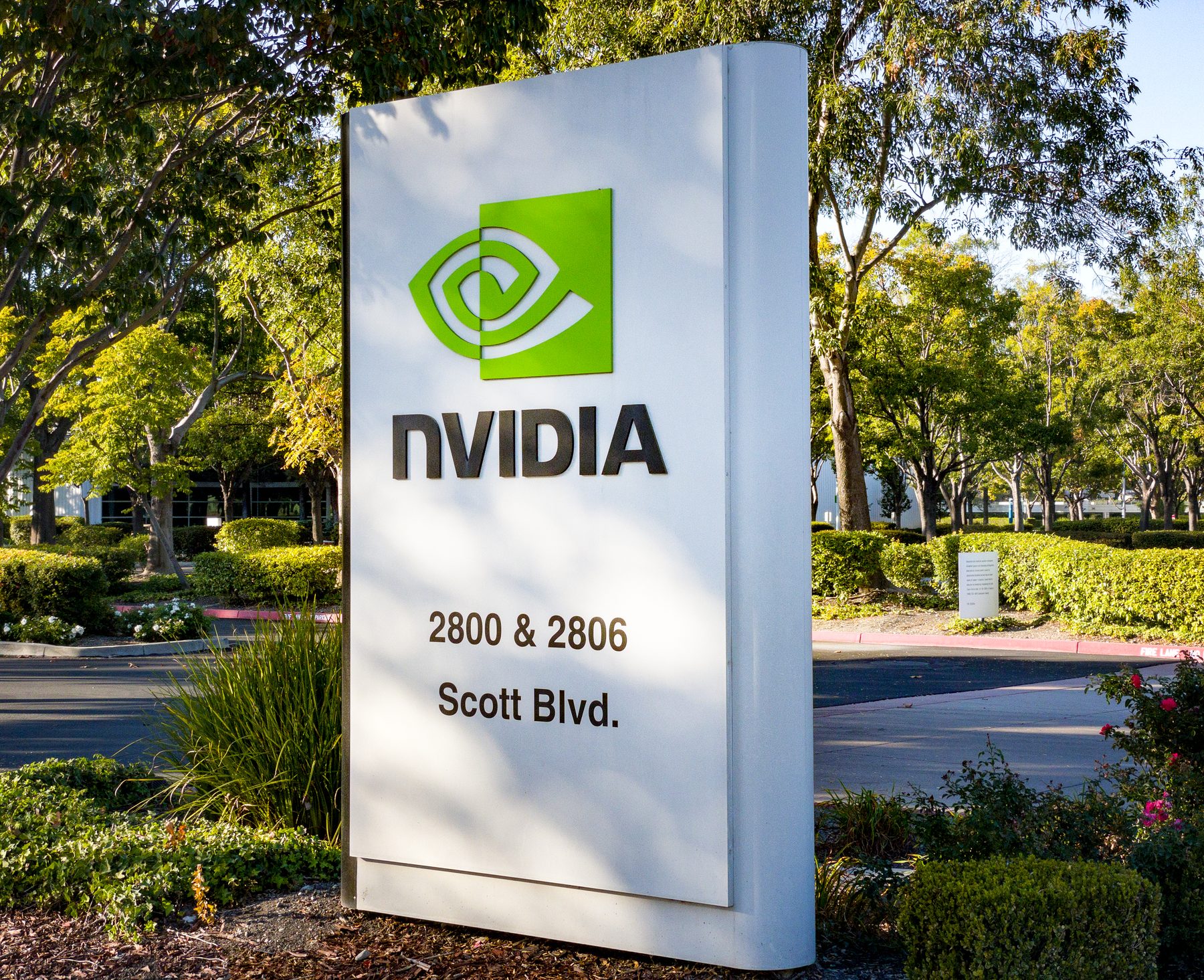 Placa preta com o símbolo verde da NVIDIA e a palavra NVIDIA em branco, apoiada em pedras cinza, com arbustos baixos e grama na frente e um prédio de vidro de janelas verticais ao fundo, sob céu claro
