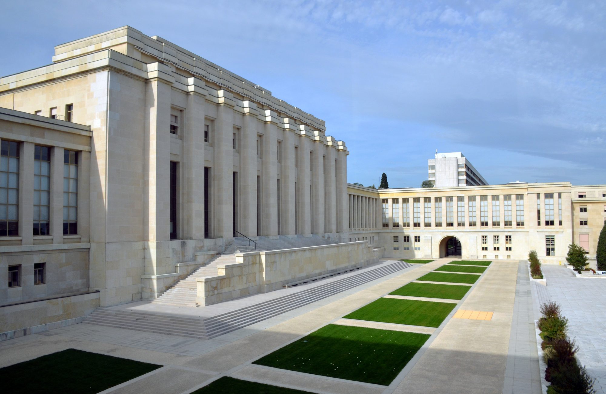 Fachada do Palais des Nations, sede da ONU em Genebra: prédio de pedra clara com colunas altas, escadaria lateral e pátio com canteiros retangulares de grama, sob céu azul