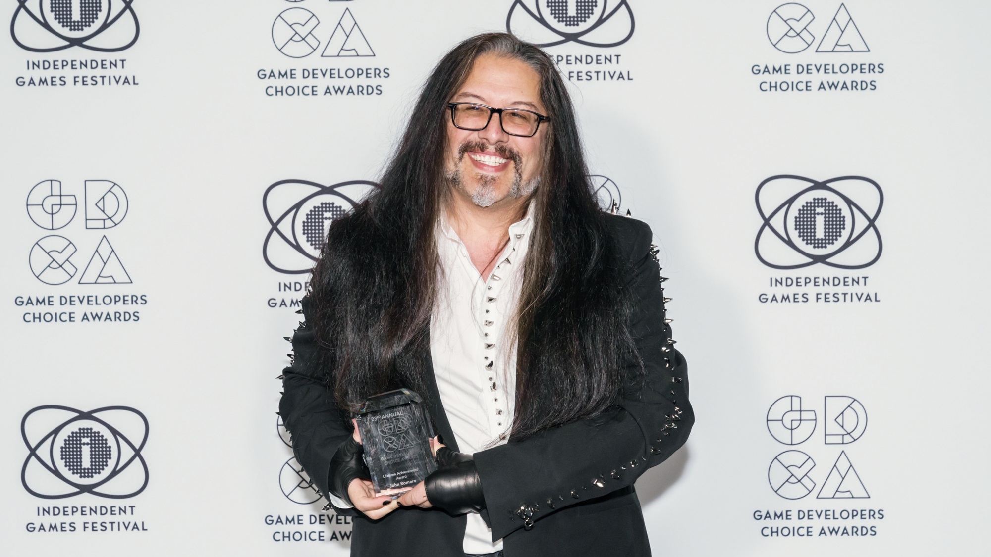 John Romero sorrindo, de óculos, cabelo comprido, camisa branca e casaco preto, segurando troféu diante de painel de premiação
