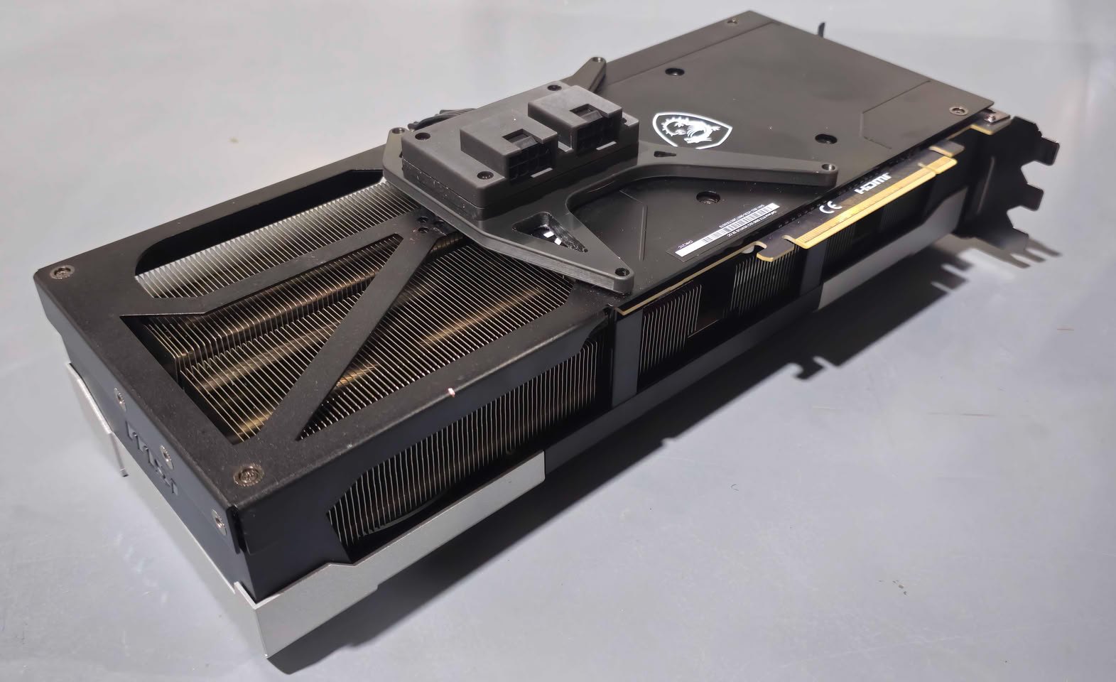 Placa de vídeo MSI GeForce RTX 5090 apoiada em uma bancada cinza exibindo um módulo preto com dois conectores de alimentação de oito pinos instalado sobre o backplate