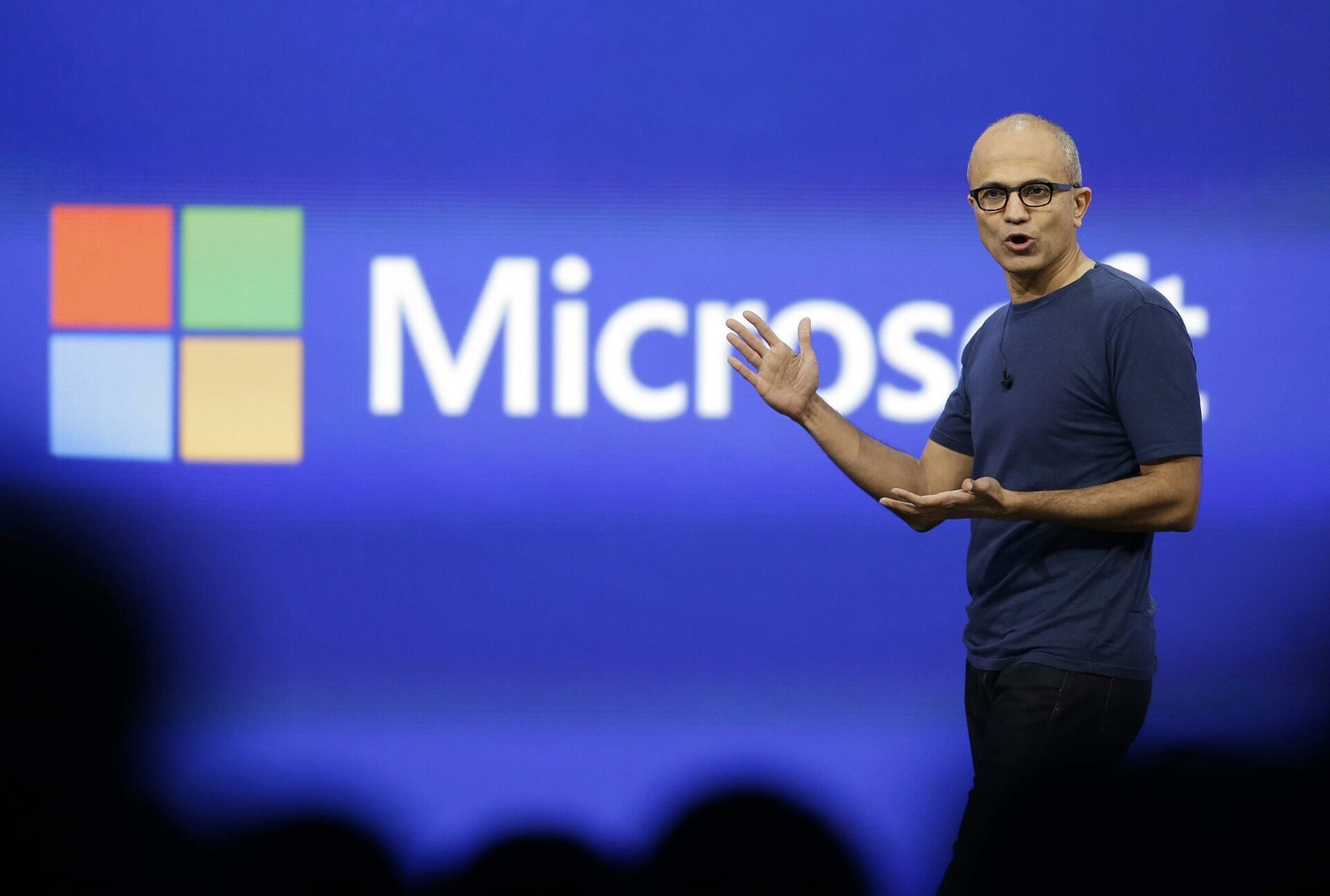 Satya Nadella vestindo camiseta azul escura e óculos, com os braços abertos gesticulando no palco diante de um telão azul com o logotipo da Microsoft