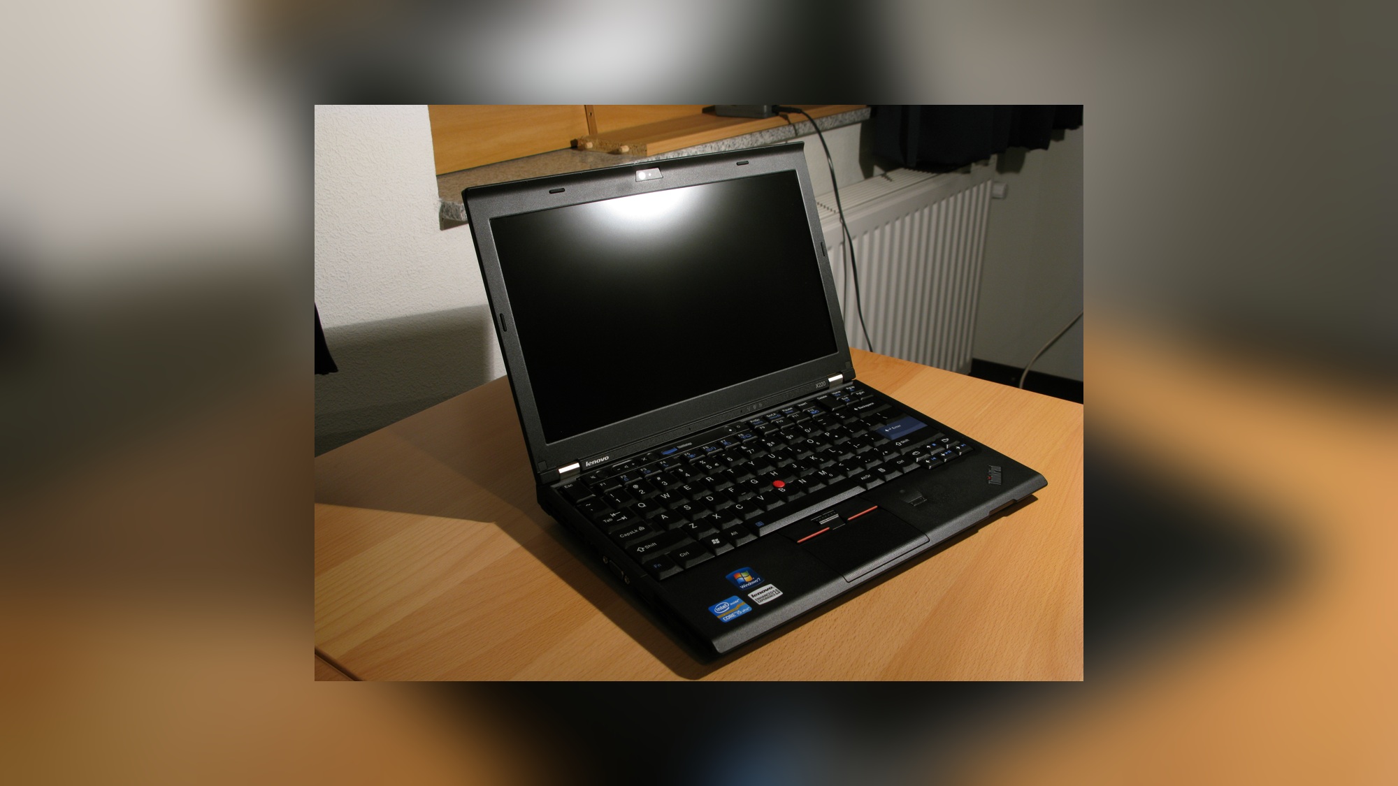 Foto ilustrativa de notebook preto ThinkPad aberto sobre mesa de madeira, com tela escura, teclado com botão vermelho e radiador branco ao fundo; não mostra o Sculpt OS