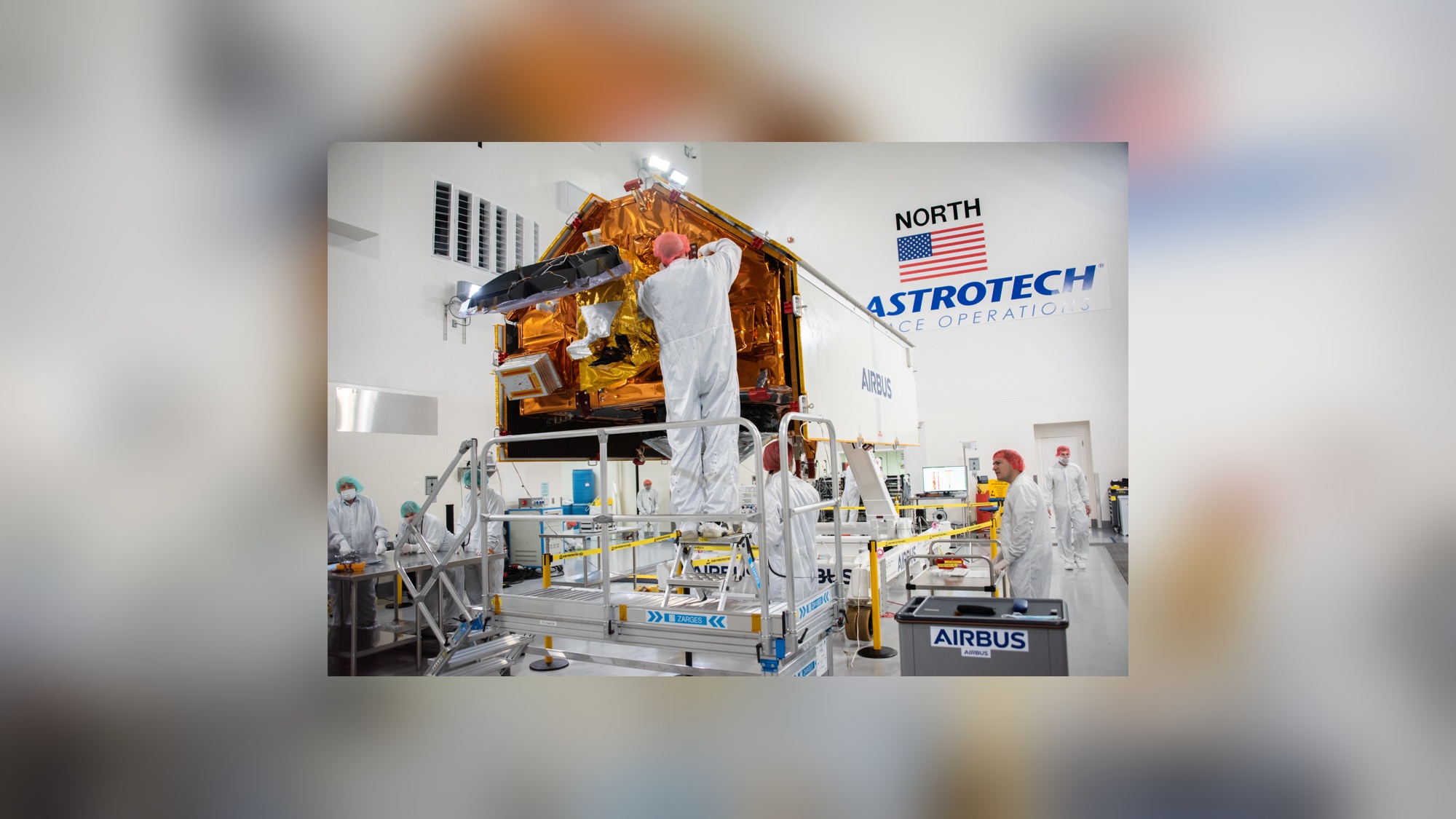 Técnicos com roupas brancas e toucas trabalham ao redor de satélite revestido de material dourado, em sala com plataformas metálicas, bandeira dos Estados Unidos e inscrições Astrotech e Airbus