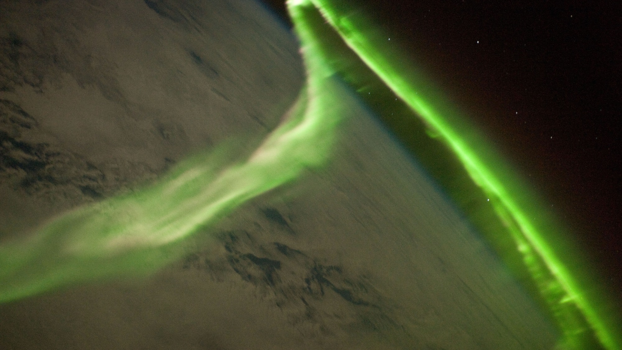Faixas luminosas verdes sobre nuvens e a curvatura da Terra, com fundo escuro e pequenos pontos de luz